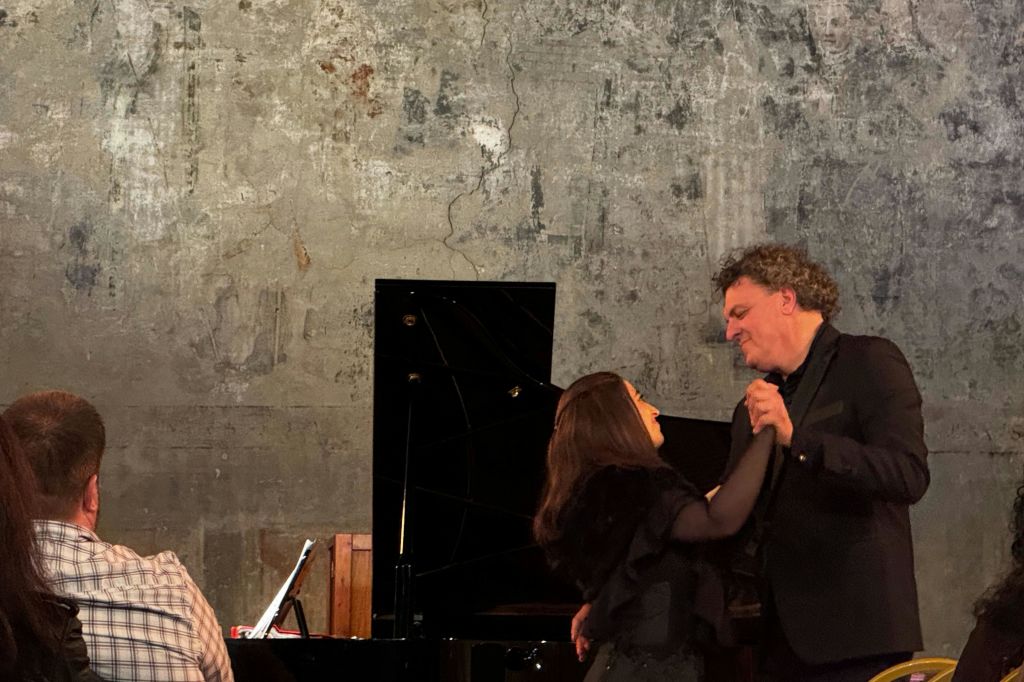 남녀가 질감이 살아있는 벽을 배경으로 그랜드 피아노 앞에서 춤을 추고 있습니다.