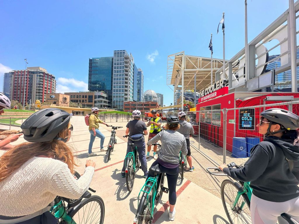 한 무리의 자전거 타는 사람들이 도시 광장에서 가이드의 말을 듣고 높은 건물과 붉은 양보가 맑은 하늘 아래 서 있습니다.