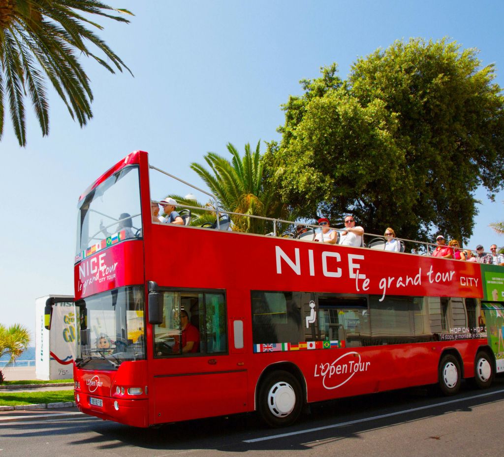 "Nice, le grand tour" 라고 표시된 빨간색 2층 투어 버스가 야자수 근처에 주차된 오픈탑 데크에 승객들과 함께 있습니다.