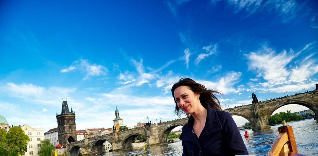 검은 외투를 입은 여성이 유서 깊은 돌다리와 푸른 하늘을 배경으로 강가에서 미소를 짓고 있습니다.