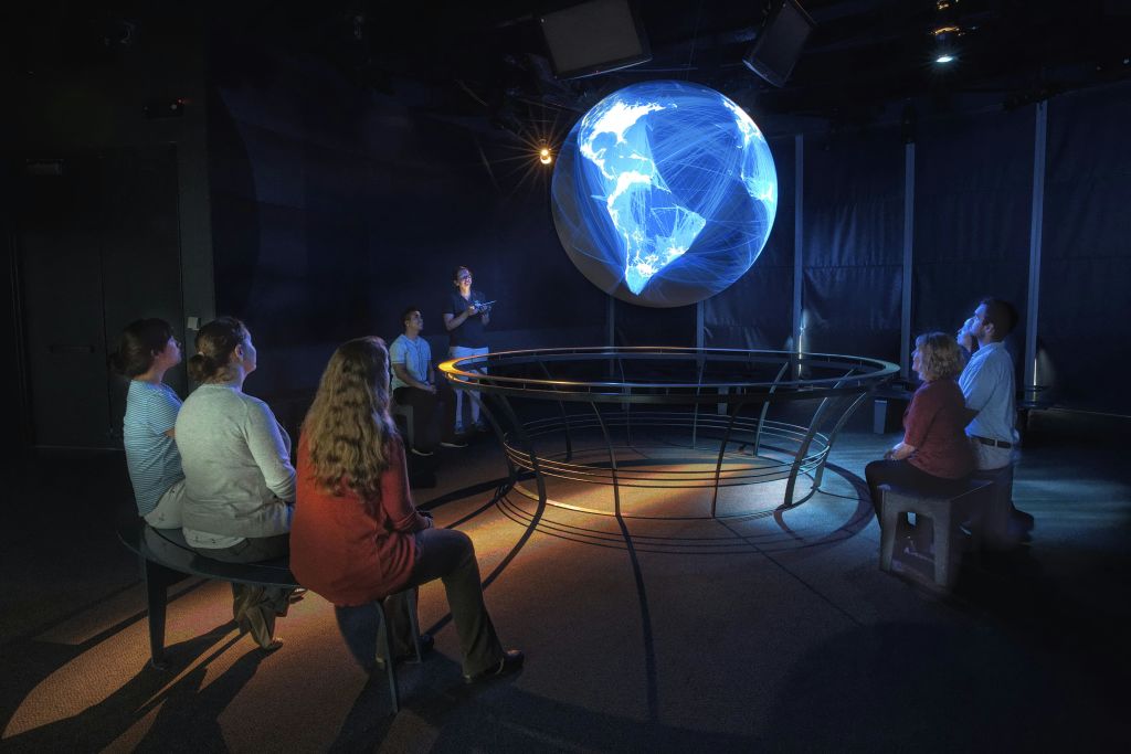 사람들은 어두운 방에 앉아 대륙 간 연결을 보여주는 네트워크 라인이 있는 조명이 켜진 지구본 디스플레이를 보고 있습니다.