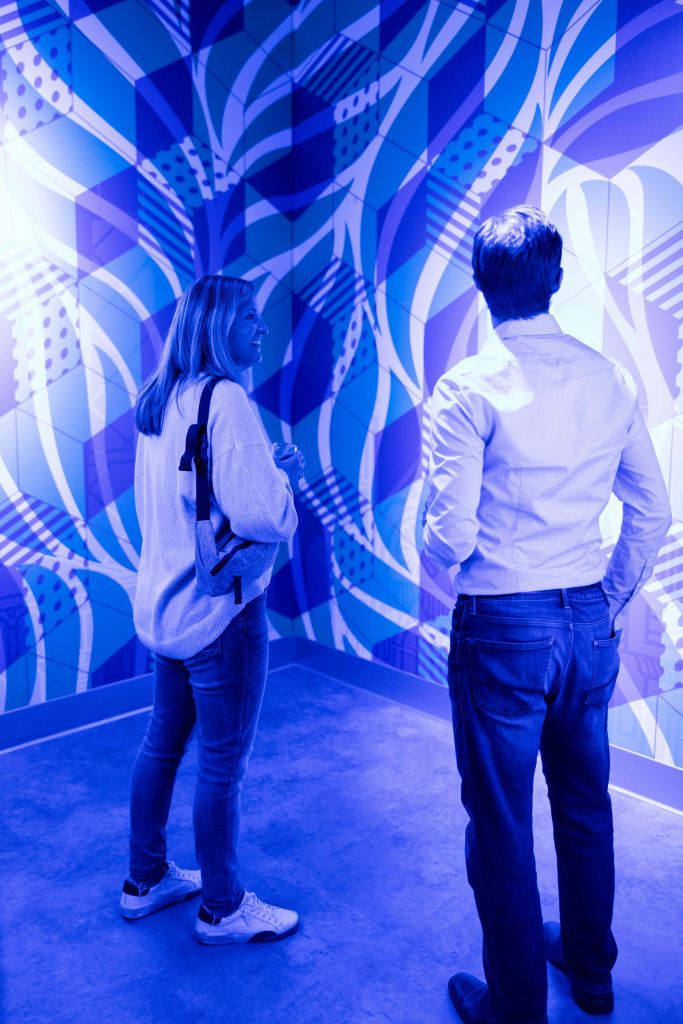 파란색 패턴의 벽이 있는 방에 두 사람이 서서 추상적인 예술 작품을 감상하며 대화를 나누고 있습니다.