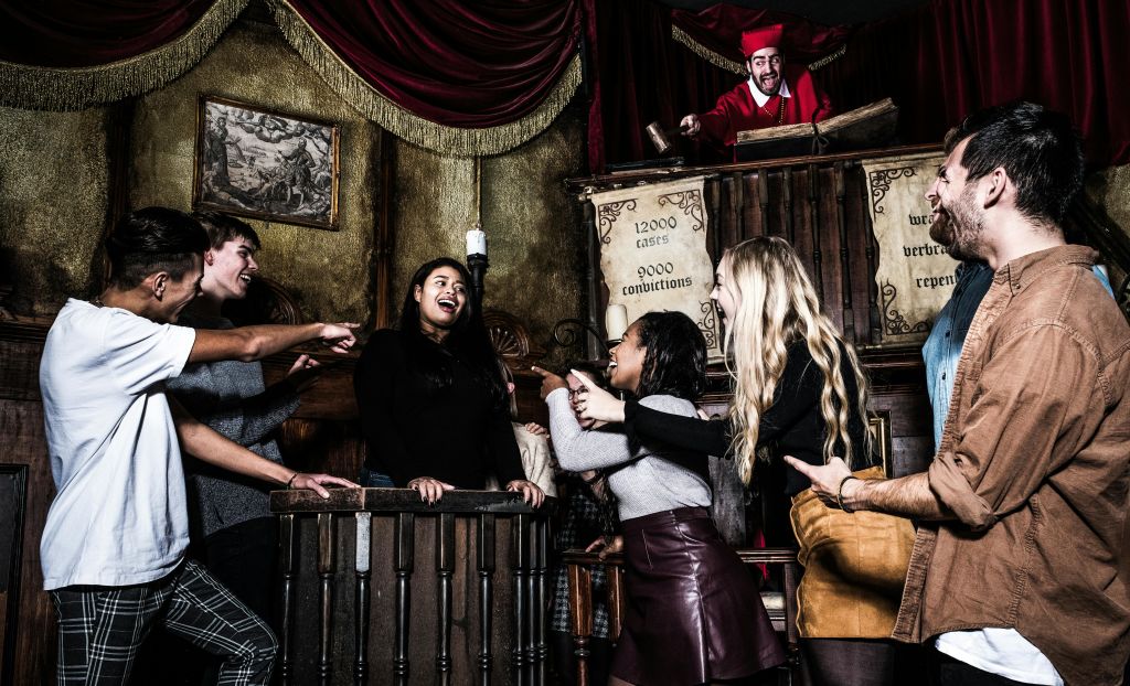 오래된 법정에서 여러 사람이 미소를 지으며 판사와 함께 나무 스탠드에 서 있는 여성을 가리키고 있습니다.