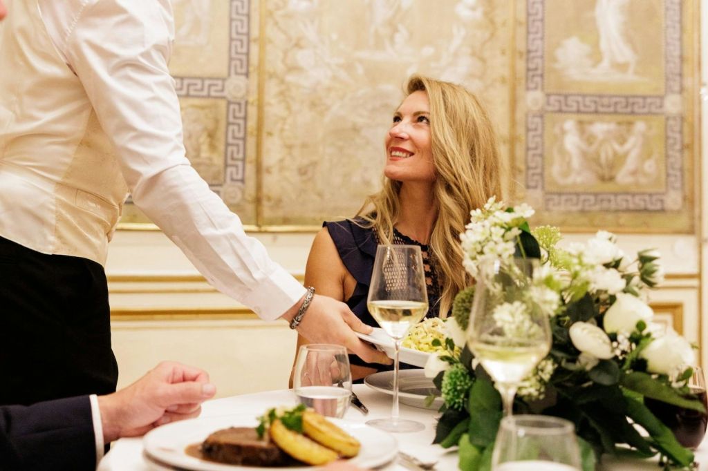 화이트 플라워와 와인잔이 놓인 우아한 식탁에서 한 여성이 웨이터를 보며 미소를 짓고 있습니다.