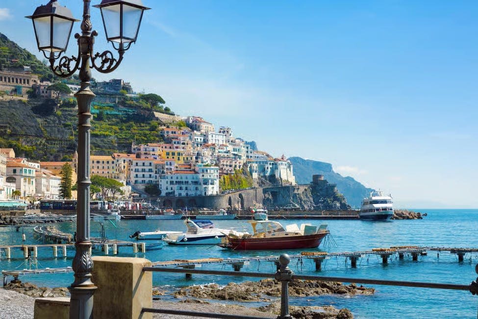 맑고 푸른 하늘이 펼쳐진 언덕길에서 물속의 보트와 다채로운 건물이 있는 항구 풍경. 전경의 램프 포스트.