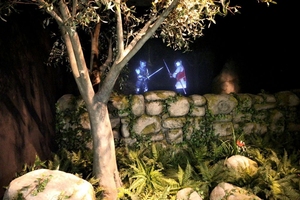 어두운 조명 속에서 나무와 양치류로 둘러싸인 돌담 뒤에서 칼을 든 두 명의 무장한 인물이 마주하고 있다.