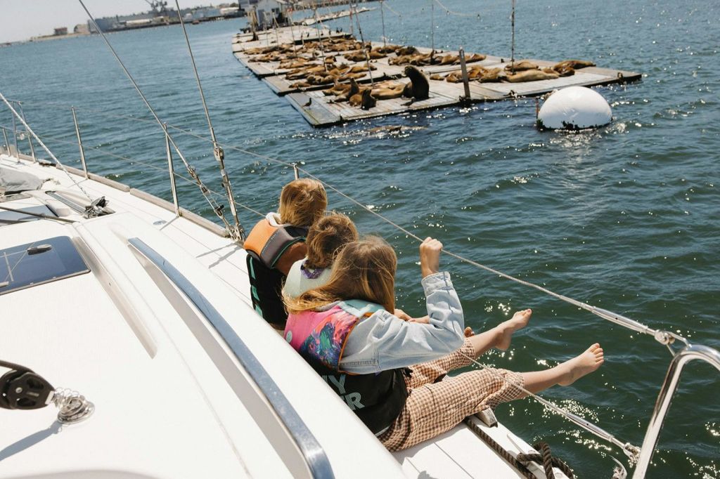 구명조끼를 입은 세 명의 어린이가 데크에 발을 걸치고 앉아 미끼 부두에 누워 있는 바다사자를 바라보고 있습니다.