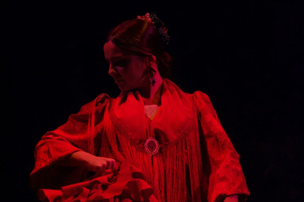 꽃무늬 헤어피스와 프린지 디테일로 장식된 빨간색 의상을 입은 여성이 저조도에서 춤을 추며 포착됩니다.