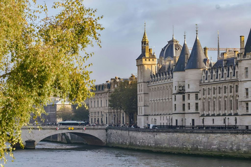 첨탑이 있는 유서 깊은 강변 건물과 전경의 나무에 둘러싸인 다리가 있습니다.
