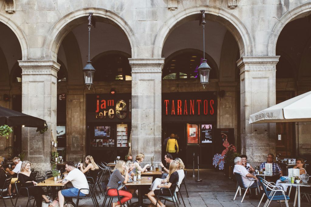 사람들이 식사하는 야외 카페 풍경. 배경에는 석조 아치와 "타란토스" 라고 적힌 팻말이 보입니다.