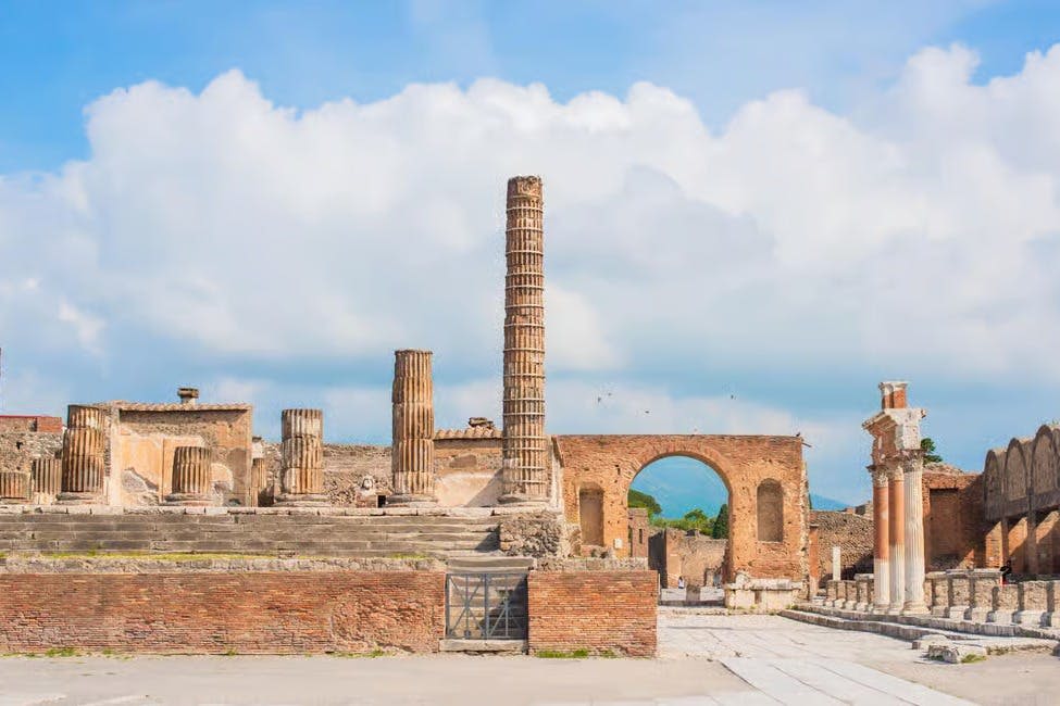 여러 개의 기둥, 석조 구조물, 부분적으로 흐린 하늘을 배경으로 한 벽돌 아치형 통로가 있는 고대 유적.