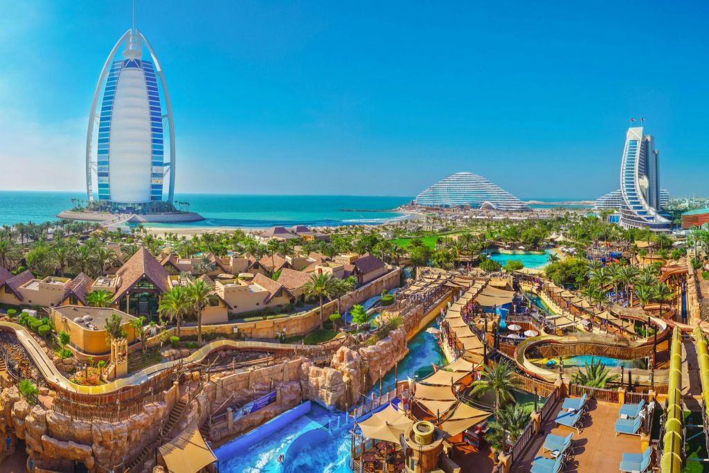 바다 근처에 미끄럼틀, 수영장, 야자수가 있는 활기찬 워터파크. 현대적인 건물이 배경으로 보입니다.