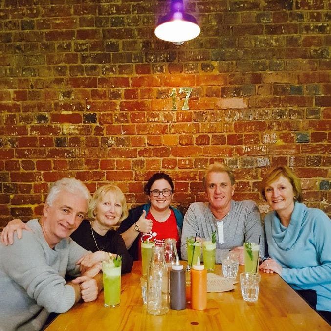 벽돌 벽에 걸린 나무 테이블에 다섯 명이 앉아 미소를 짓고, 테이블 위에 초록색 음료와 양념이 놓여 있습니다.