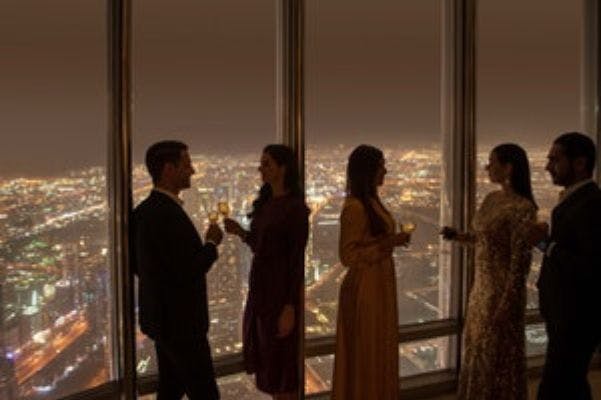 우아하게 차려입은 네 사람이 커다란 창가에 서서 밤에는 도시 풍경을 배경으로 한 음료를 들고 있습니다.