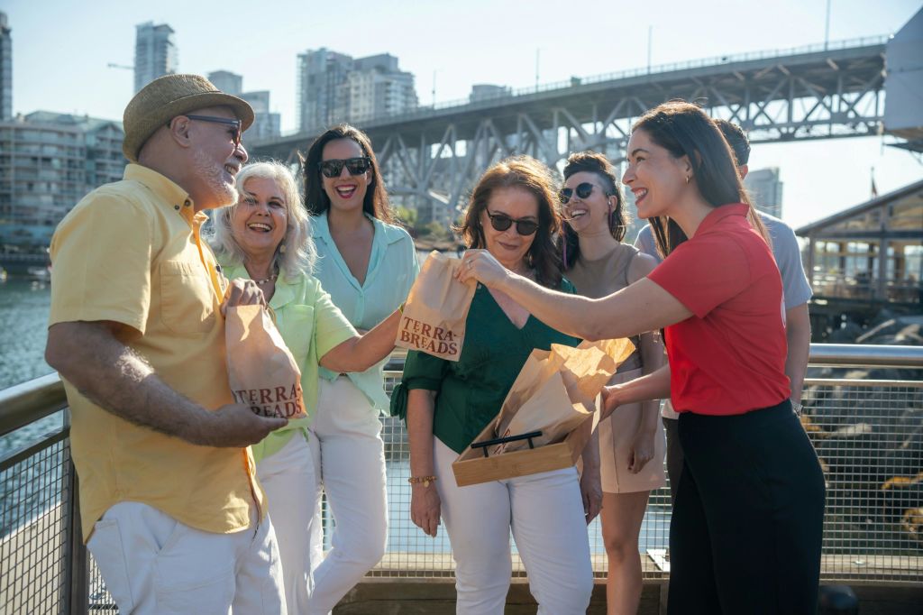 다리와 도시 건물이 배경으로 놓인 수변 난간 옆에서 5명이 테라 브레드 가방을 들고 미소를 짓고 있습니다.