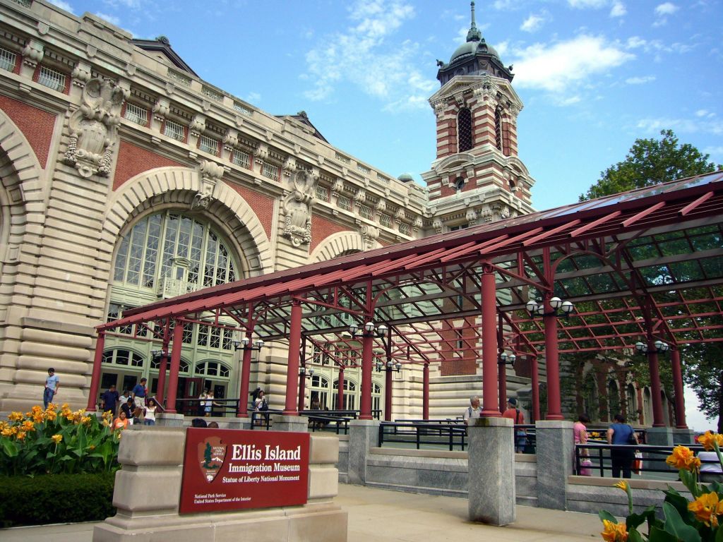 화려한 외관, 붉은 캐노피 구조, 외부 방문객이 있는 엘리스 섬 이민 박물관 건물.