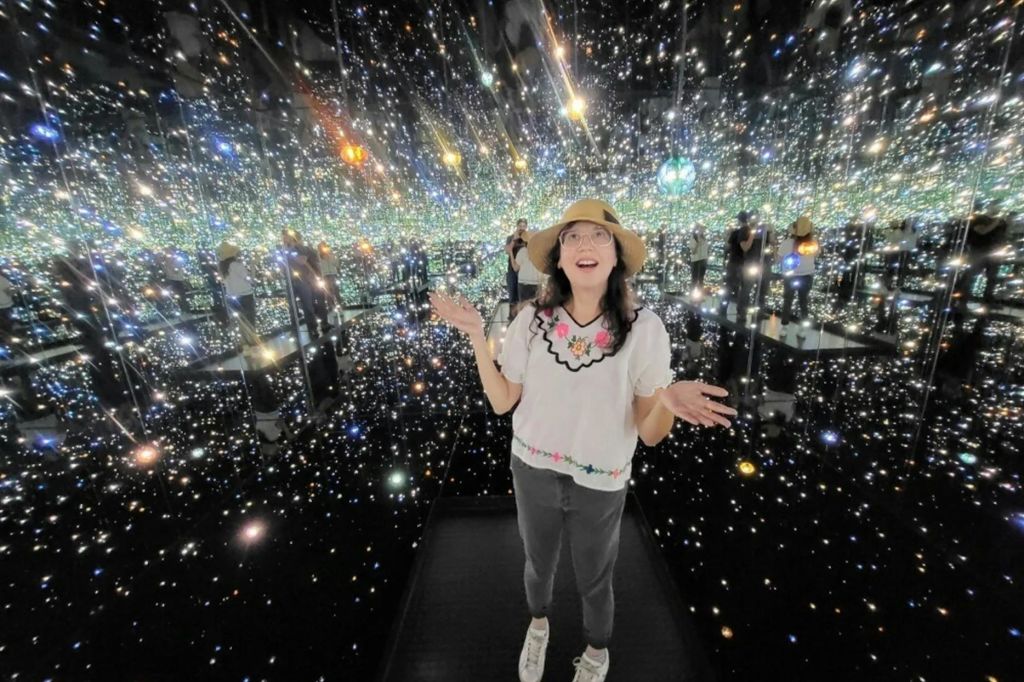 거울 벽과 반짝이는 불빛이 가득한 방에 한 여성이 모자, 안경, 꽃무늬 상의, 회색 바지, 운동화를 신고 서 있습니다.