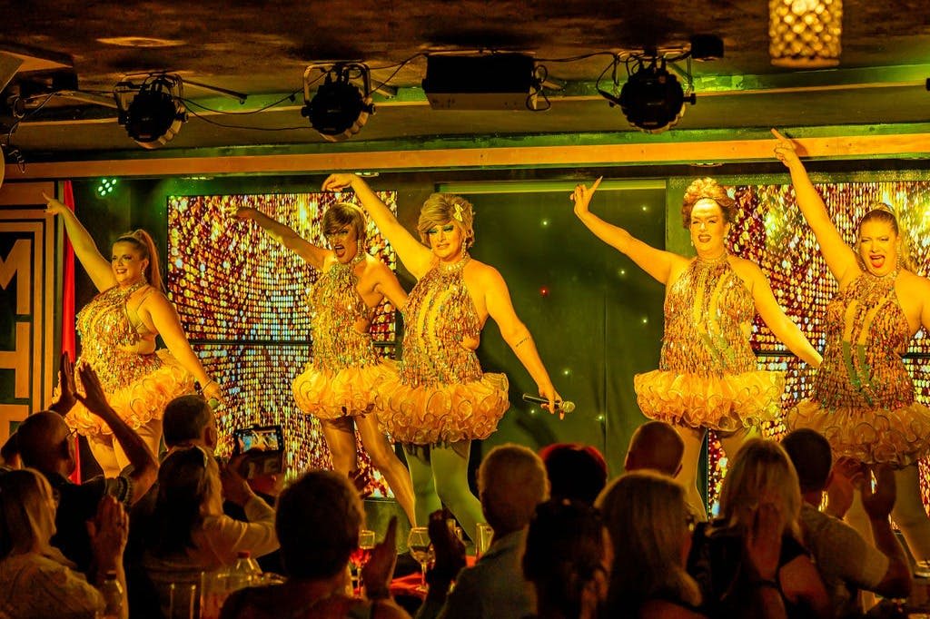 화려한 주황색 의상을 입은 네 명의 공연자가 무대에서 활기찬 조명 아래 관객들이 박수를 치며 춤을 추고 있습니다.