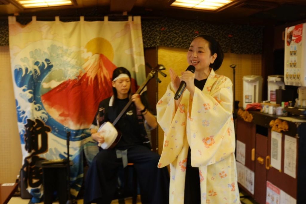 노란색 기모노를 입은 여성이 앉아 있는 사람이 전통 현악기를 연주하는 동안 마이크를 불렀습니다. 그 뒤에는 산의 배경이 있습니다.