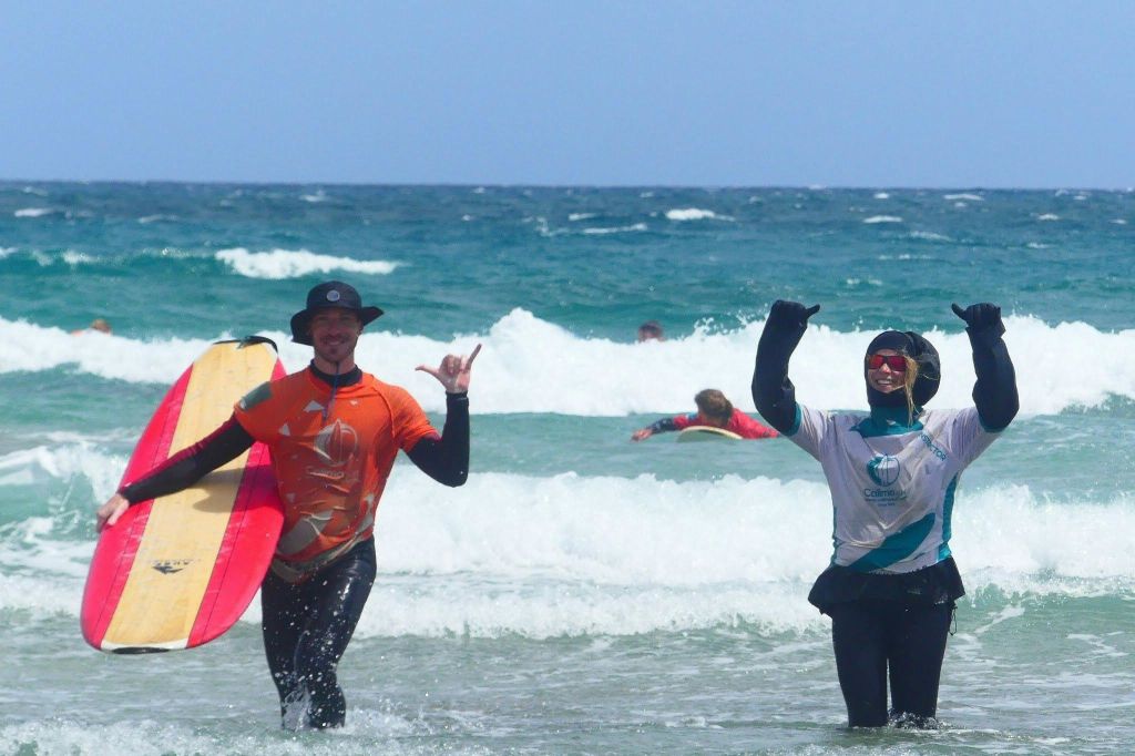 오렌지 셔츠와 검은 모자를 쓴 사람이 서핑 보드를 들고 제스처를 취합니다. 배경에는 다른 서퍼들이 있습니다.