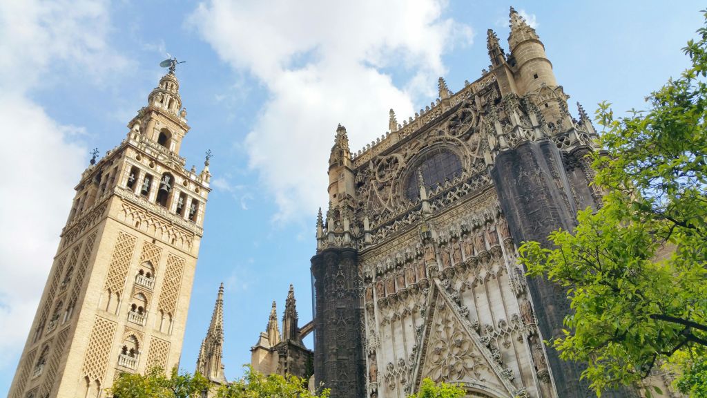 화려한 외관과 왼쪽에 높이 솟은 종탑이 있는 고딕 양식의 성당으로, 구름이 흩어져 있는 푸른 하늘과 맞닿아 있습니다. 녹색 단풍이 눈앞에 펼쳐집니다.