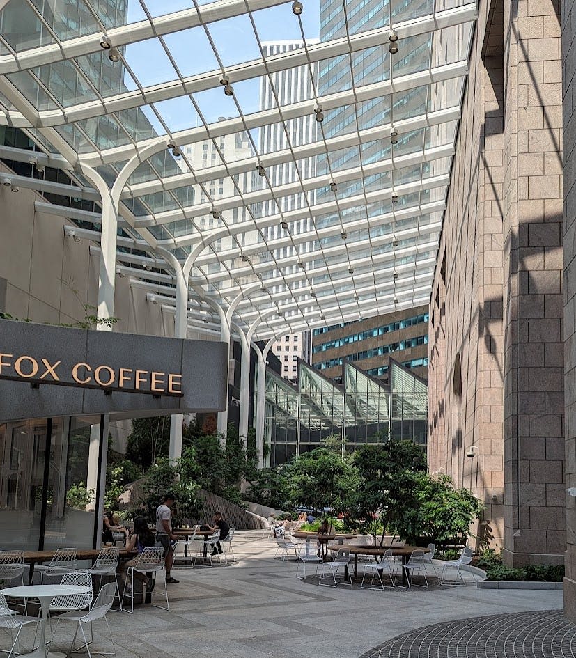 '폭스 커피' 간판이 있는 건물과 인접한 유리 지붕으로 덮인 테이블, 의자, 식물이 있는 야외 광장입니다.