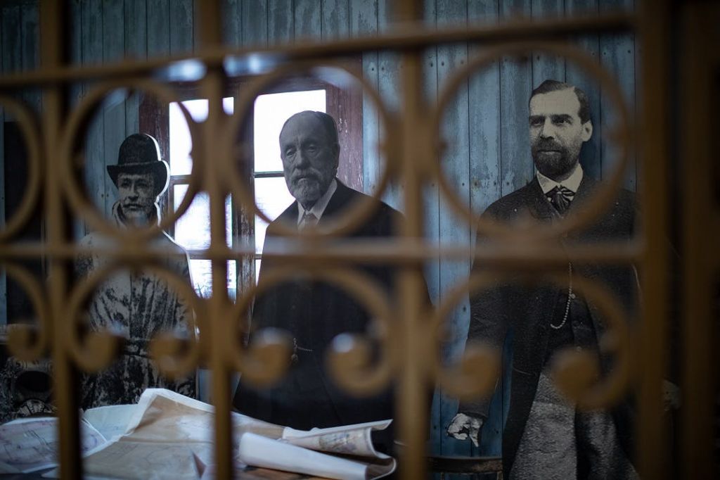 장식용 철공 뒤에 정장을 입은 남성의 역사적인 흑백 초상화 두 장. 롤업된 문서가 전경에 표시됩니다.