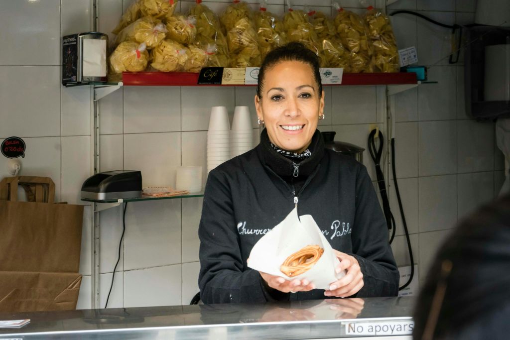 검은 재킷을 입은 여성이 카운터 뒤에 서서 미소를 짓고 말린 페이스트리를 종이에 담은 채 뒤로 간식과 컵을 들고 있습니다.