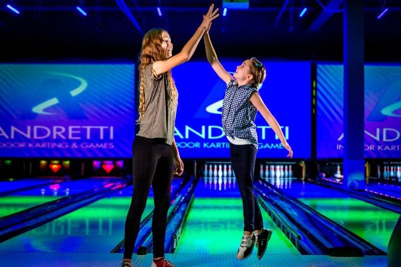 배경 화면에 "Andretti Indoor Karting & Games" 가 보이는 활기찬 볼링 레인에서 하이파이브하는 두 사람.