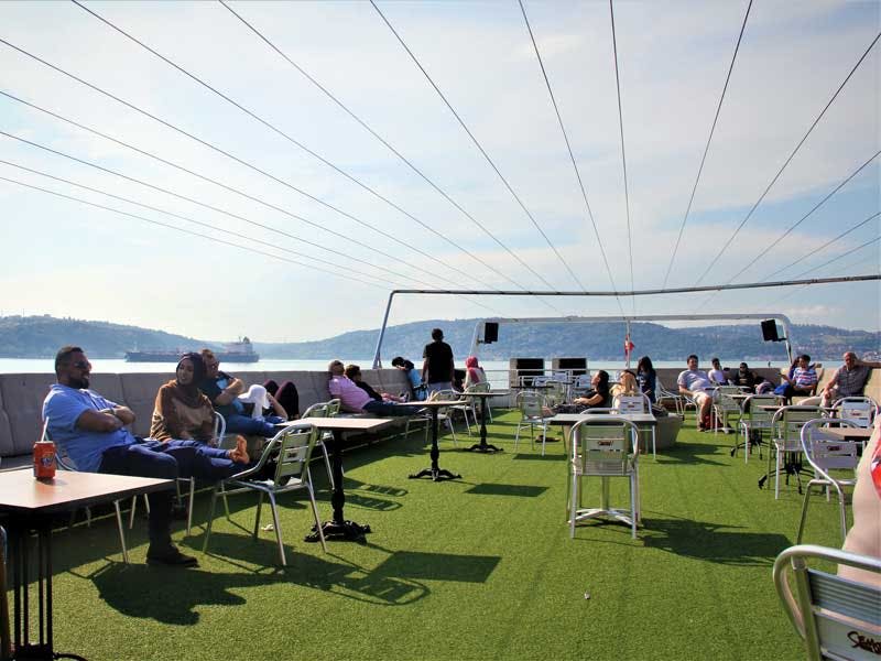 녹색 인조 잔디가 깔린 옥상 테라스에 앉아 휴식을 취하는 사람들로, 물과 먼 언덕이 내려다보입니다.