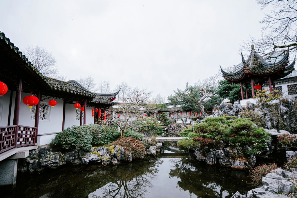 붉은 등불, 곡선 지붕, 바위가 있는 연못, 흐린 하늘 아래 다양한 나무와 관목이 있는 전통 정원.