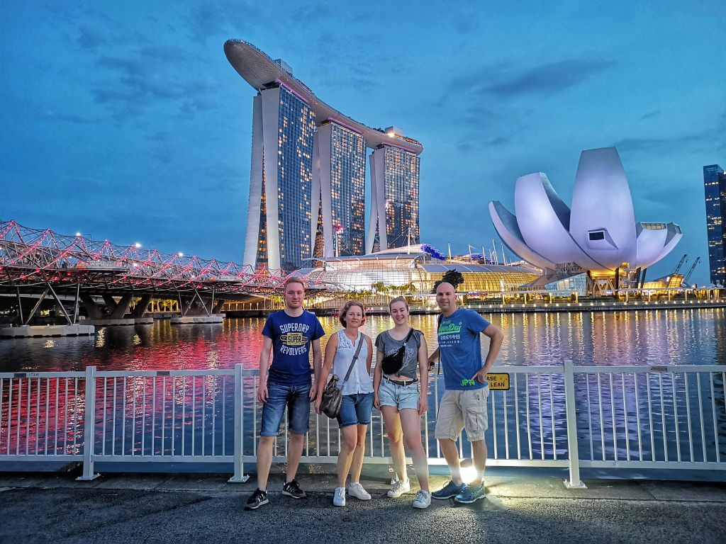 싱가포르의 마리나 베이 샌즈와 헬릭스 브리지 앞에서 4명이 포즈를 취하고 있으며, 해질 무렵에는 해안가 전망을 감상할 수 있습니다.