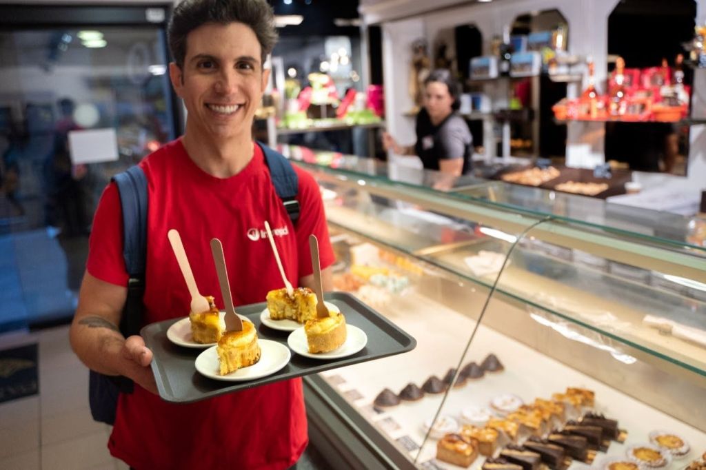빨간 셔츠를 입은 사람이 베이커리 안에 케이크 조각 세 접시가 놓인 트레이를 들고 카메라를 보며 미소를 짓고 있습니다.