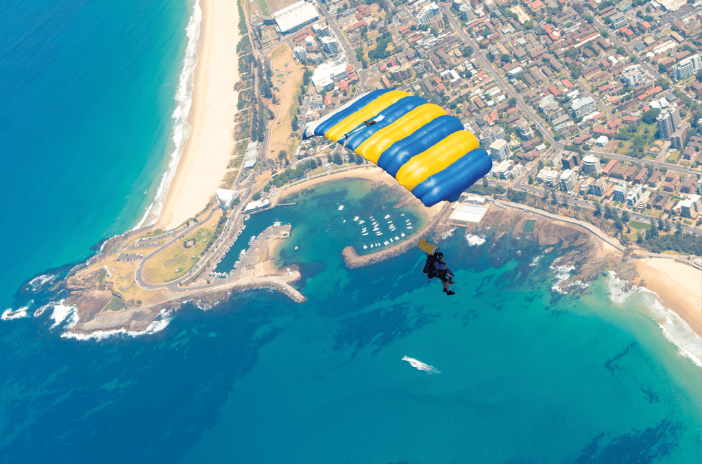 해안 도시 위로 노란색과 파란색 낙하산을 타고 내려오는 스카이다이버는 해변, 정박지, 도시 풍경을 보여줍니다.