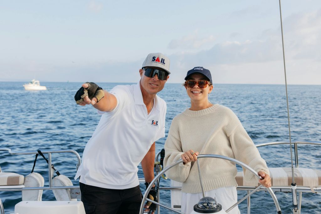 항해 중인 샌디에이고 선장이 먼 곳을 가리키고 있는 동안, 한 손님이 보트의 조종석 뒤에 서서 미소를 짓고 있습니다.