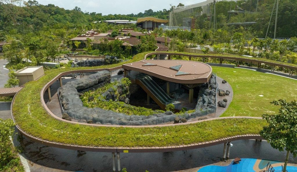 녹지, 통로 및 인근 구조물로 둘러싸인 갈색 지붕의 대형 현대식 건물이 있는 공원의 공중 전망.