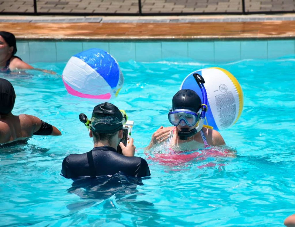 스노클링 장비를 착용하고 근처에 비치볼이 떠 있는 수영장의 두 사람. 한 명은 수영모를 쓰고 있고, 둘 다 대화에 참여하고 있는 것 같습니다.