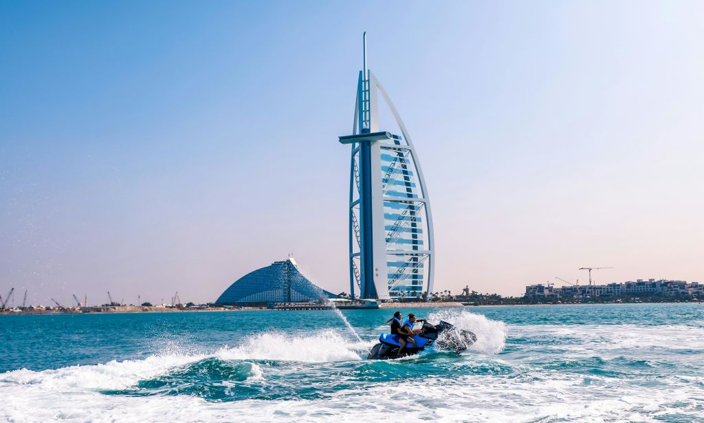푸른 바다에서 제트 스키를 타는 두 사람, 버즈 알 아랍 (Burj Al Arab) 호텔과 맑은 날을 배경으로 한 커브드 빌딩을 만나보세요.