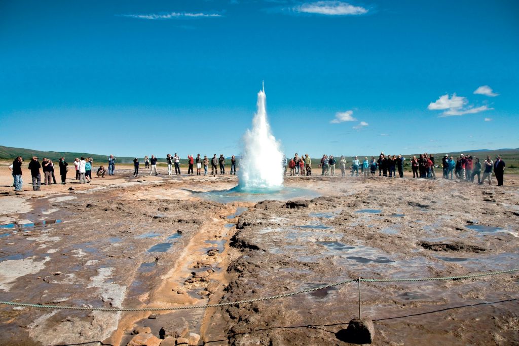 청명한 푸른 하늘 아래에서 분출하는 간헐천을 배경으로 바위 지형과 드문드문 녹음으로 둘러싸인 간헐천을 관찰하는 사람들.