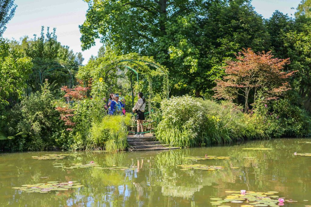꽃이 만발한 식물과 나무로 둘러싸인 울창하고 푸른 정원의 연못 옆 작은 나무 플랫폼에 두 사람이 서서 이야기를 나누고 있습니다.