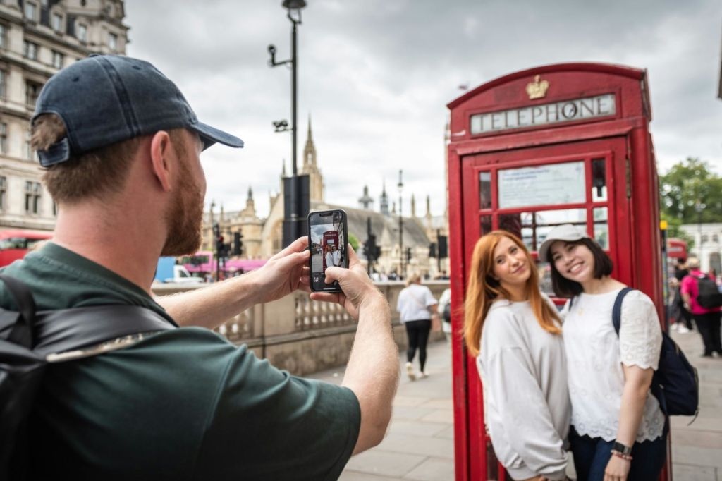 한 도시의 빨간색 전화 부스 앞에서 스마트폰으로 사진을 찍는 두 사람이 포즈를 취하고 있습니다.