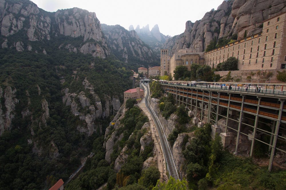 울창한 녹음과 바위 절벽이 있는 산악 지대를 가로지르는 기차 트랙. 오른쪽에 오래된 건물 단지가 보입니다.