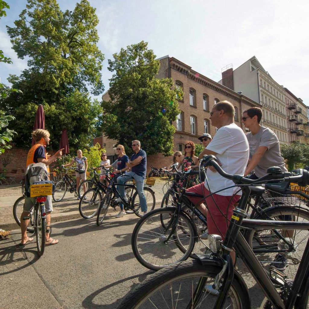 자전거를 탄 한 무리의 사람들이 벽돌 건물과 커다란 나무 근처에서 야외로 모여 대화를 나누거나 경청합니다.