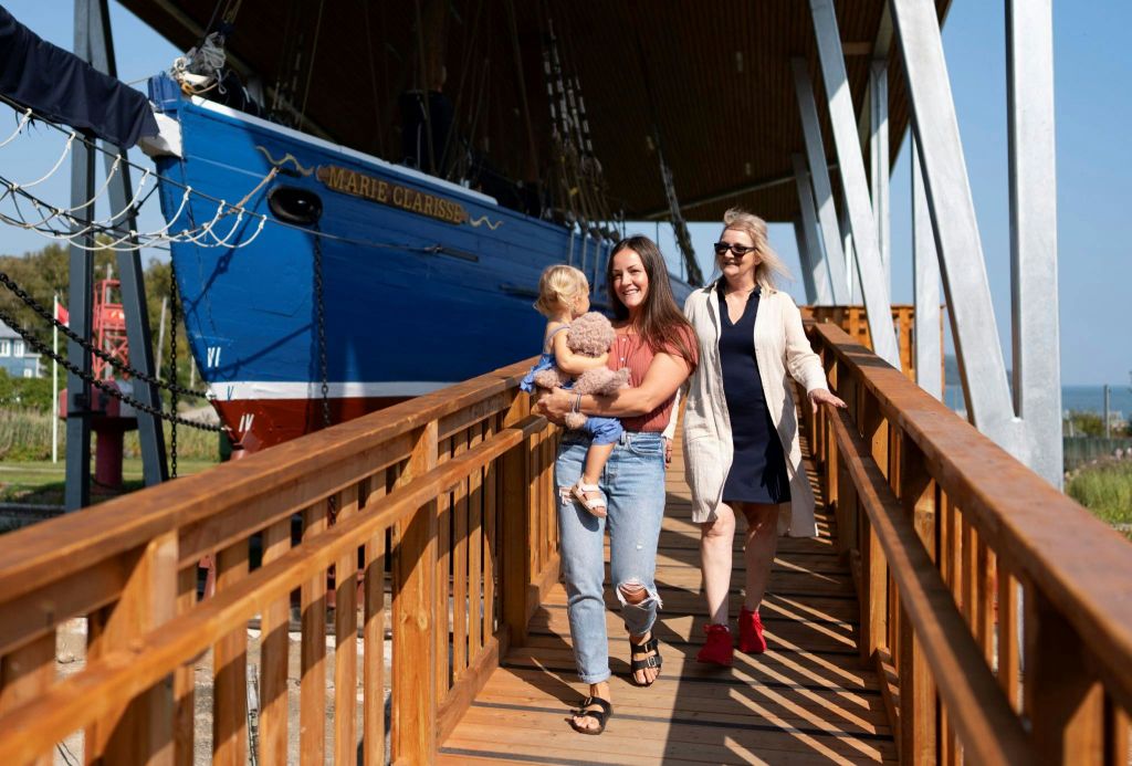 한 명은 유아를 안고 있는 두 명의 여성이 "마리 클라리스" 라는 이름의 커다란 푸른 배 옆의 나무 산책로를 걷고 있습니다.