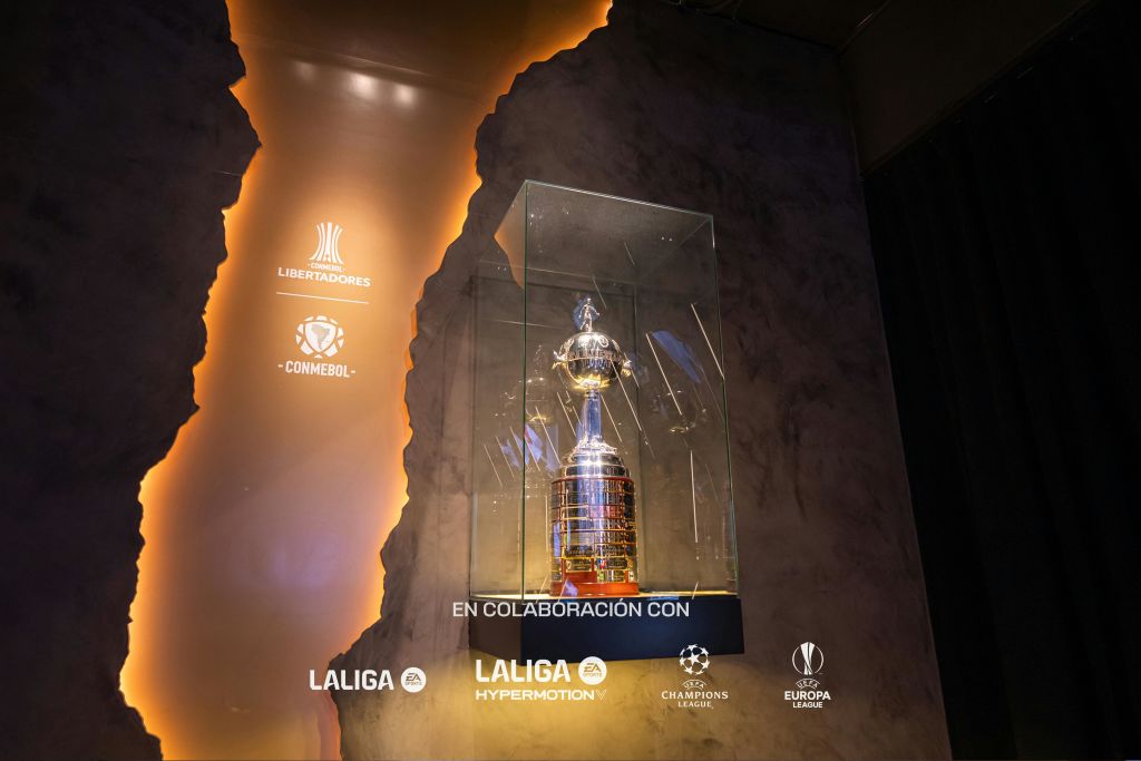 다양한 축구 리그 로고와 어두운 질감의 배경에 "EN colaboración CON" 이라는 텍스트가 있는 유리 케이스의 실버 트로피.