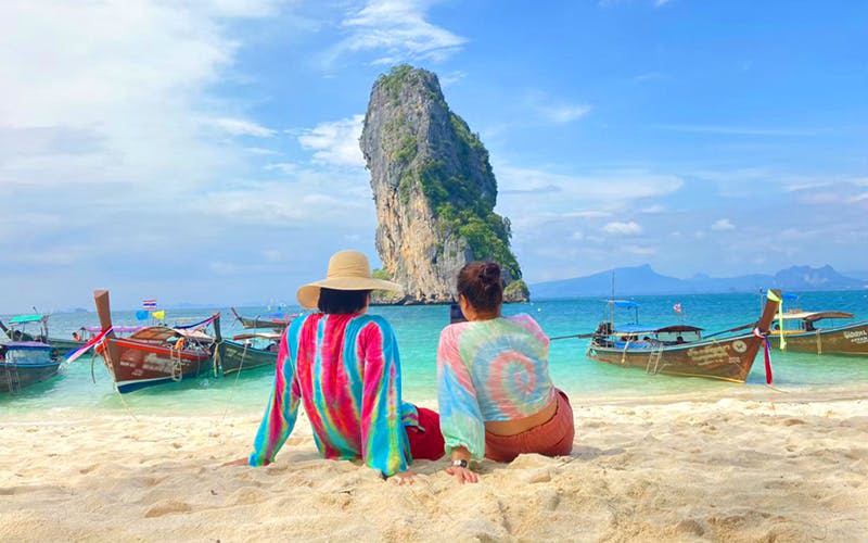 알록달록한 색상의 셔츠를 입은 두 사람이 바다를 마주한 모래 해변에 앉아 있으며, 배를 배경으로 높은 바위가 형성되어 있습니다.