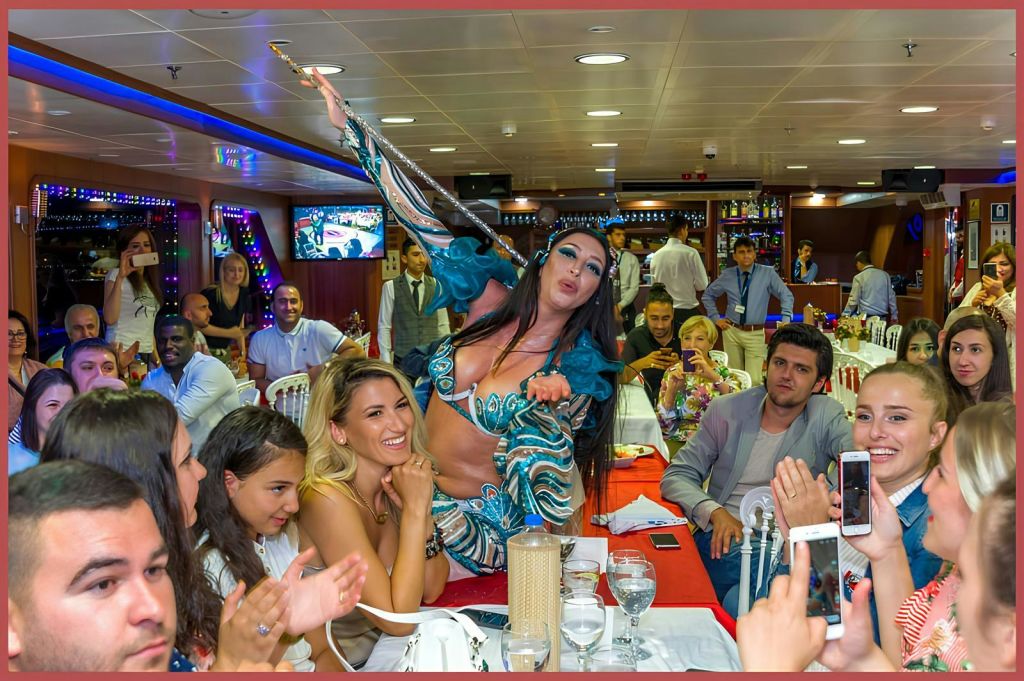 활기찬 의상을 입은 공연자가 붐비는 축제 분위기의 실내 테이블에서 춤을 추고 있습니다. 주변 사람들이 박수를 치며 사진을 찍고 있습니다.