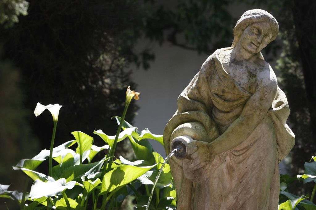 녹음이 우거진 단풍과 하얀 칼라 백합으로 둘러싸인, 물을 붓는 형상의 풍화된 석상.