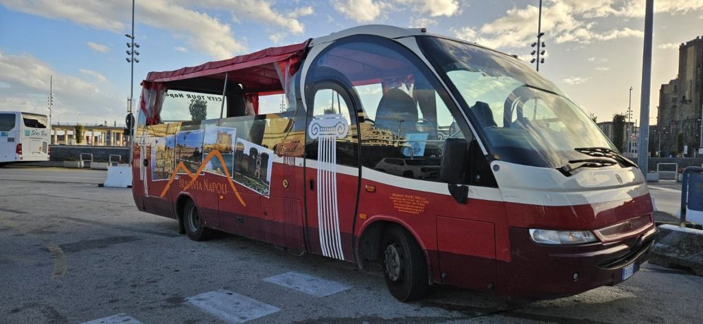 커다란 창문과 나폴리 명소 홍보 이미지가 있는 빨간색 관광 버스가 흐린 하늘 아래 많이 주차되어 있습니다.