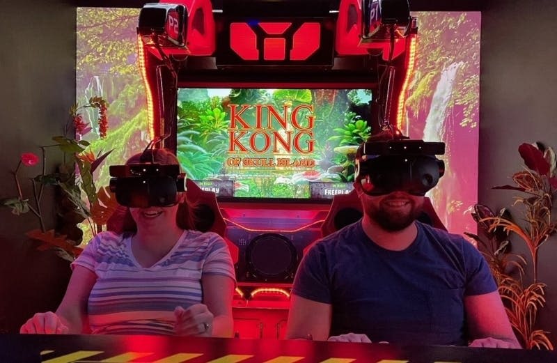 VR 헤드셋을 착용한 두 사람이 뒤쪽 스크린에 "King Kong: Of Skull Island" 가 표시된 빨간색 아케이드 좌석에 앉아 있습니다.